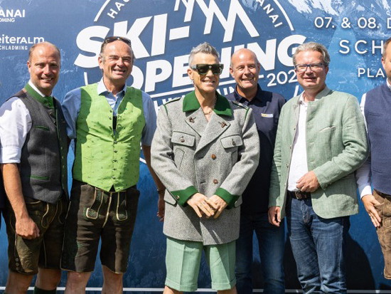 Robbie Williams (Mitte) und die Veranstalter:innen freuen sich jetzt schon auf das Event im Dezember.