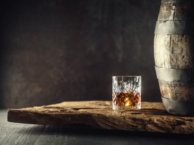 Glass of whisky cognac or bourbon in ornamental glass next to a vinatge wooden barrel on a rustic wood and dark background.