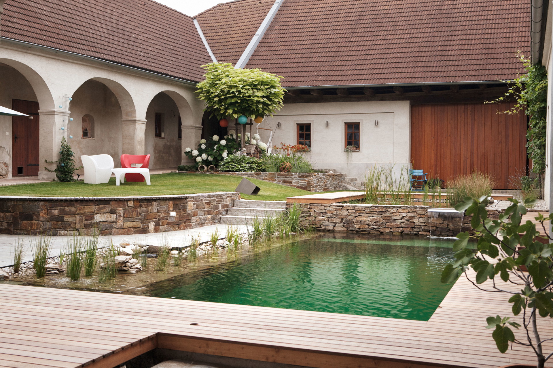 Ab Hof: Naturpool auf zwei Ebenen in einem alten Gehöft inklusive Unterwassersitzbank. Naturstein- und Holzterrassen fassen das Pool-Ensemble ein. kramerundkramer.at