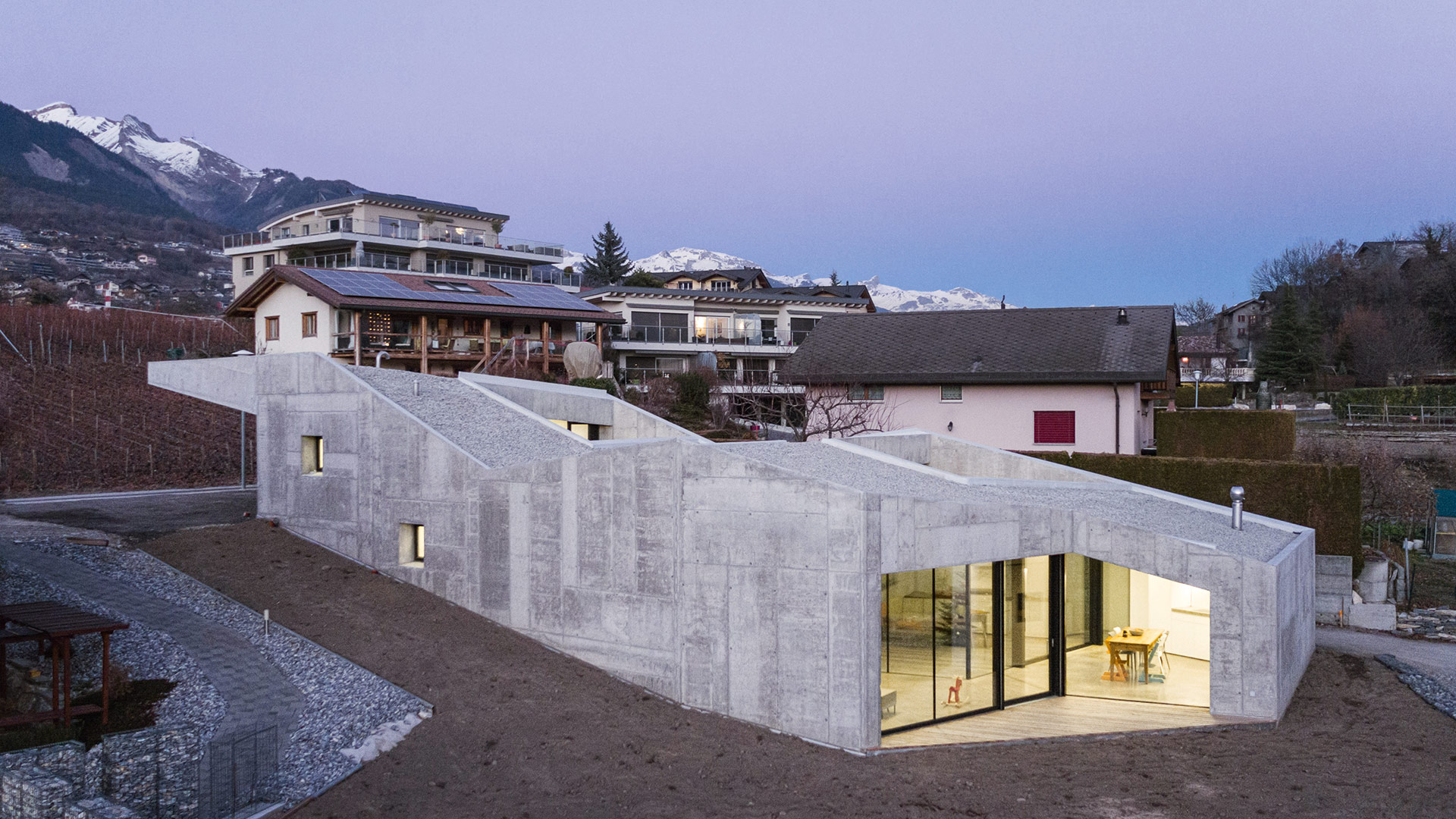 In unmittelbarer Nähe der Rhône schlummert dieses Wohnhaus im schweizerischen Savièse. Die charakteristische Dachkante spiegelt die Silhouette der Alpen wider. Innen besteht das Haus aus fünf unterschied-lichen, ineinander verschachtelten Split-Level-Wohnebenen. Durch drei kleine Patios gelangt Tageslicht ins Innere des Gebäudes. Eine Besonderheit ist die glatte, industrielle Oberfläche des Werkstoffs, die die mono-lithische Erscheinung des Hauses unterstreicht – als Schalung kamen nämlich Metallplatten zum Einsatz. anakoarchitecture.ch