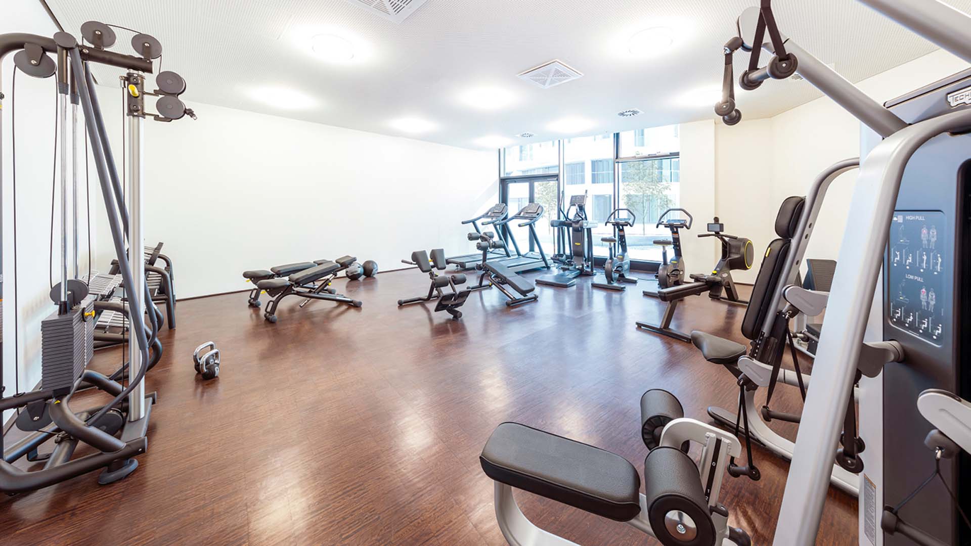 Attraktiver Fitnessraum mit TECHNOGYM®-Geräten und Blick auf die begrünte Plaza.