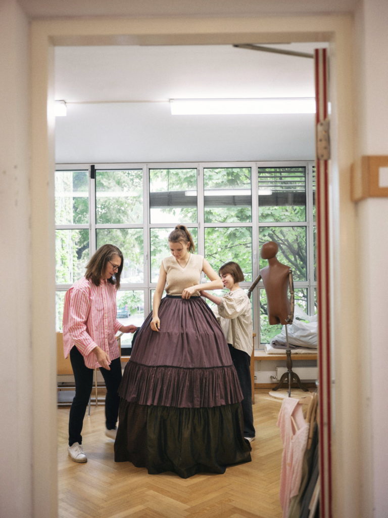 Ein Reifrock für Helena. Kostümhospitantin Paula Glawion pielt, einen Probenrock genäht. Esther Geremus und ihre Assistentin Maria-Lena Poindl helfen beim Anziehen. 
