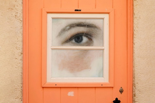 Ein gemaltes menschliches Auge blickt durch das kleine Fenster einer orangefarbenen Tür.