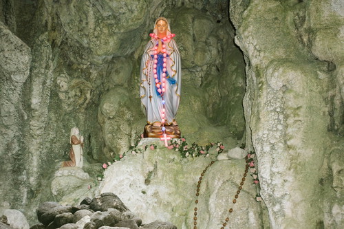 Eine bunt geschmückte Marienstatue mit Rosenkränzen steht in einer Felsgrotte, daneben eine kleinere kniende Figur.