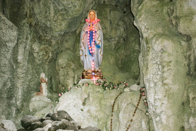 Eine bunt geschmückte Marienstatue mit Rosenkränzen steht in einer Felsgrotte, daneben eine kleinere kniende Figur.