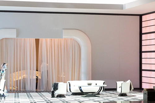 Eine elegant eingerichtete Bühne zeigt ein modernes Zimmer mit weißen Möbeln, rosa Wandelementen und einer Person im farbenfrohen Gewand, die sich streckt.