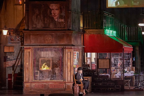 Eine Szene aus einem Theaterstück, die einen stilisierten Cafégarten zeigt. Ein älterer Mann sitzt auf einem Stuhl, umgeben von nostalgischen Details wie Werbeschildern und einer Telefonzelle. Die Beleuchtung erzeugt eine dramatische Atmosphäre, die die Melancholie des Ortes unterstreicht.