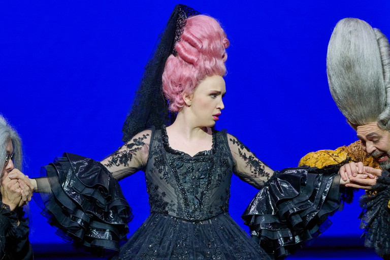 Eine Frau mit hochgesteckter rosa Perücke und schwarzer Spitze trägt ein ausladendes schwarzes Kleid vor tiefblauem Hintergrund und blickt energisch zur Seite.