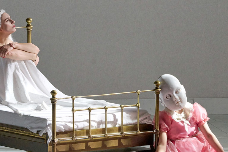 Am Fußende eines Metallbetts sitzt eine unheimlich wirkende Puppe mit Maske und rosa Kleid auf dem Boden