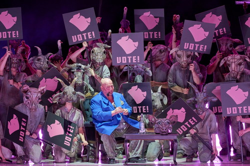 Ein Mann im blauen Sakko sitzt vor einer Gruppe maskierter Figuren, die rosa Schilder mit der Aufschrift „VOTE!“ und einem Schweinekopf-Symbol hochhalten.