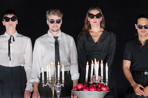 Personen in Schwarz-Weiß-Kleidung und Sonnenbrillen stehen ernst nebeneinander hinter Kerzenleuchtern und einer Schale mit roten Äpfeln.
